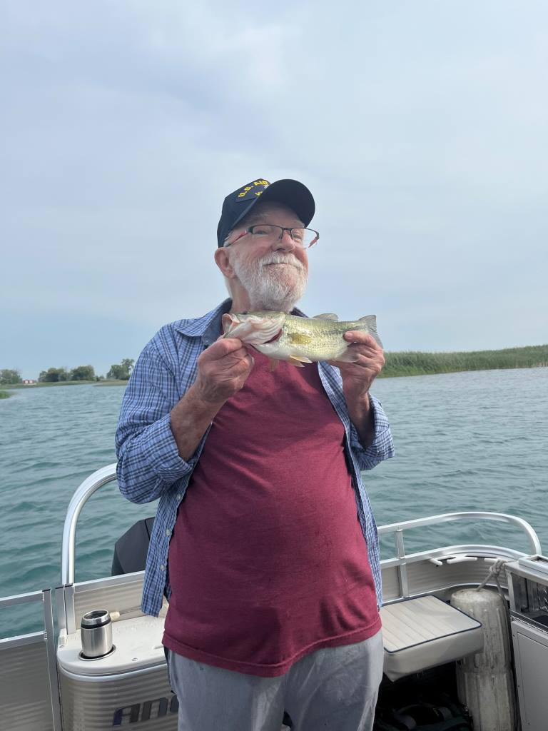 Man on a boat holding a small fish, smiling. Overcast sky, water in the background.