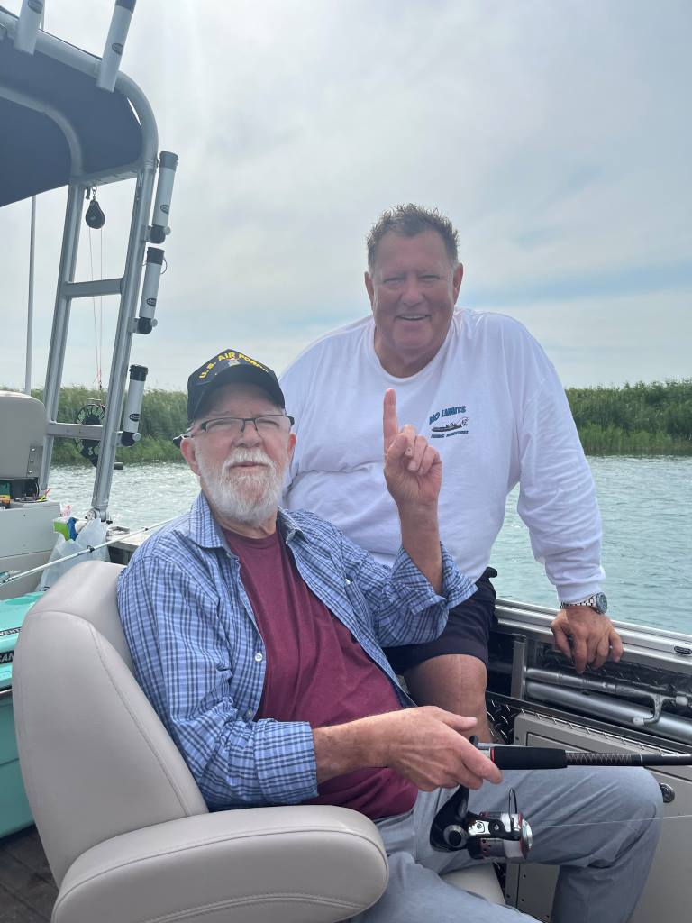 Two men fishing on a boat; one points up with a fishing rod in hand.