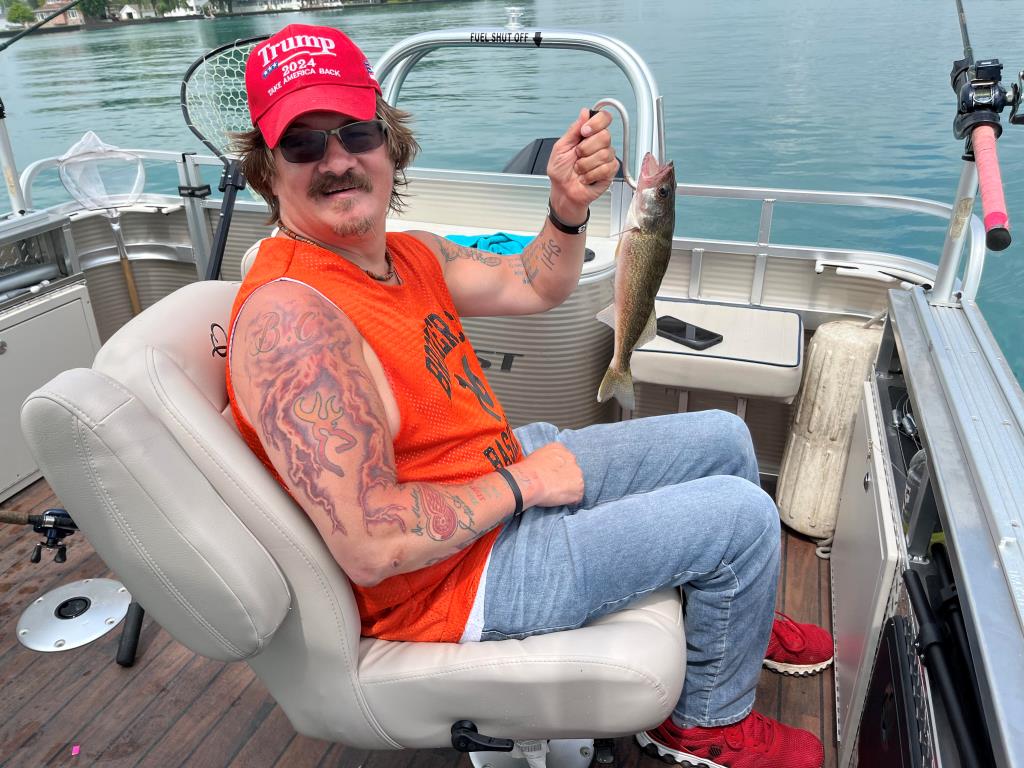 Man on boat, holding fish. Wearing hat and orange shirt, smiling. Lake background.