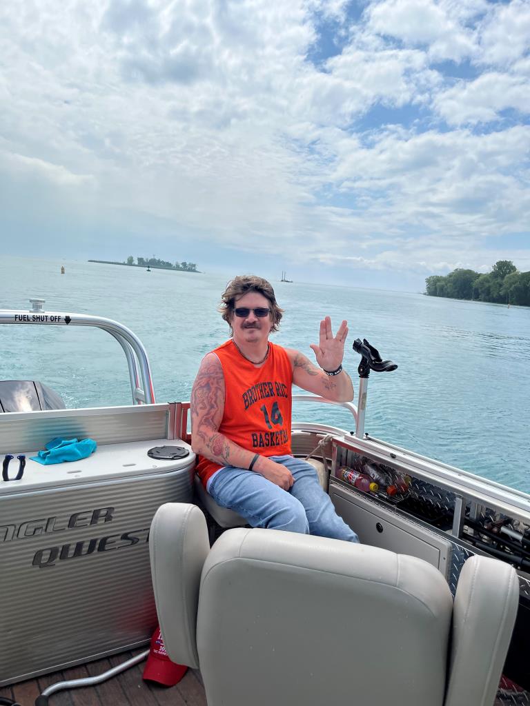 Man waving on a pontoon boat, blue water and sky in the background.