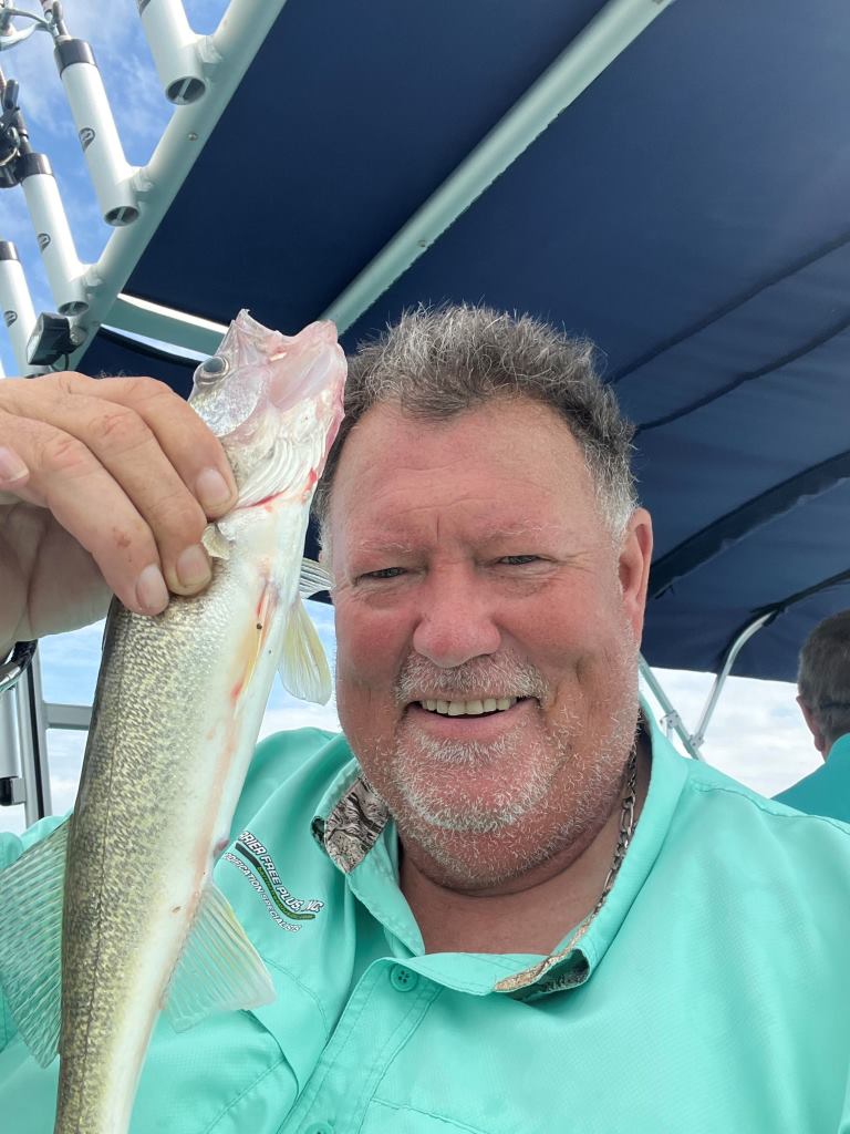 Man on boat holding up fish, smiling, wearing turquoise shirt, sunny day.