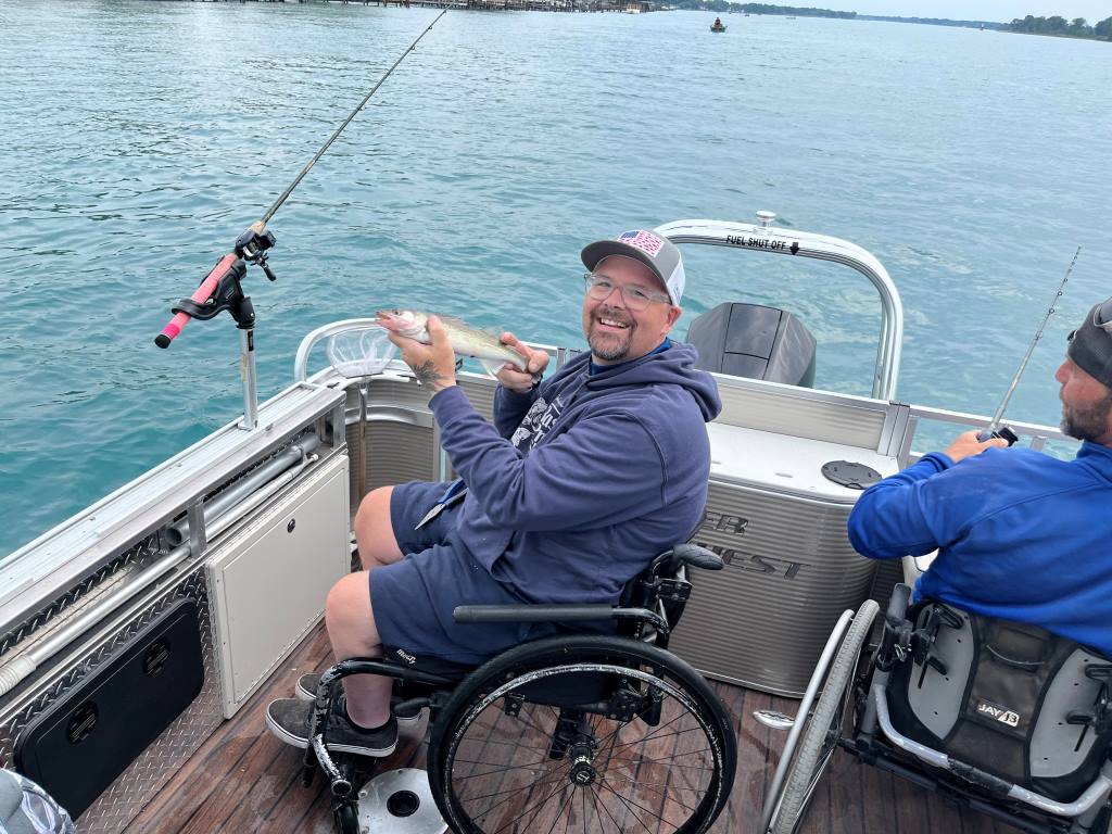 Man in wheelchair on a boat, holding a fish, smiling. Another man fishes nearby. Blue water setting.