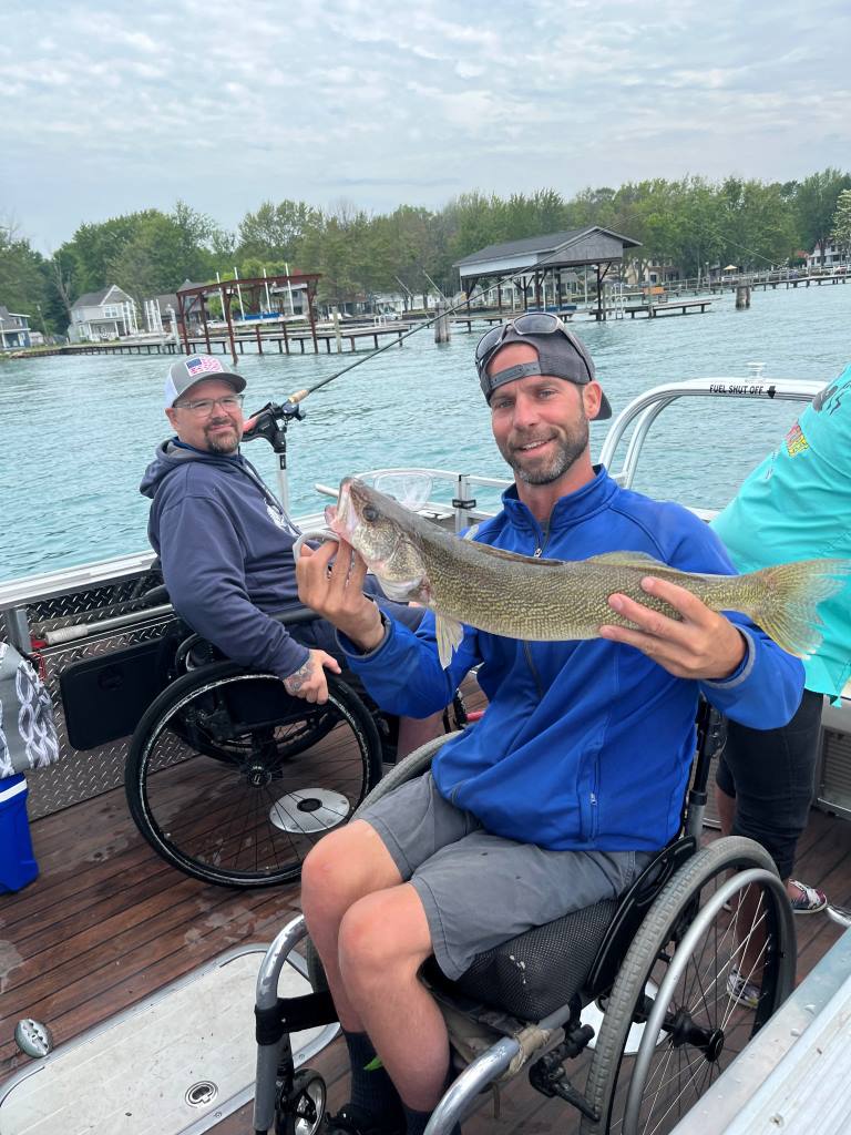 Two men in wheelchairs on a boat fishing. One holds up a walleye. Blue water and sky.