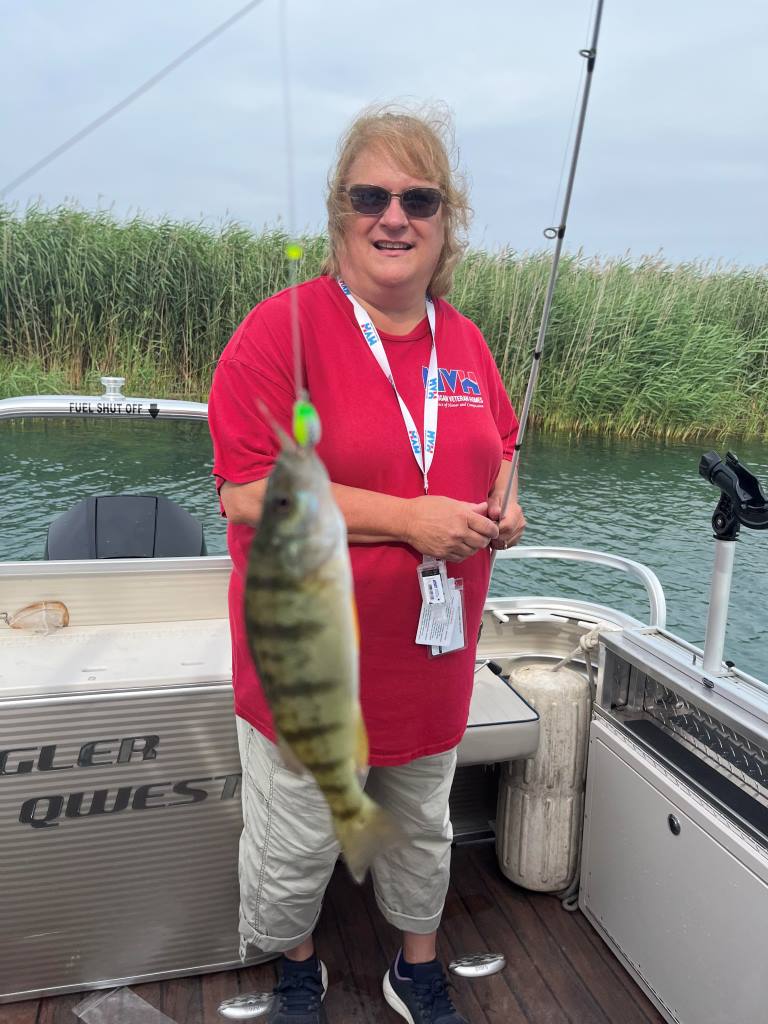 Woman on a boat, holding a yellow perch she caught, with fishing rod and tall reeds in background.
