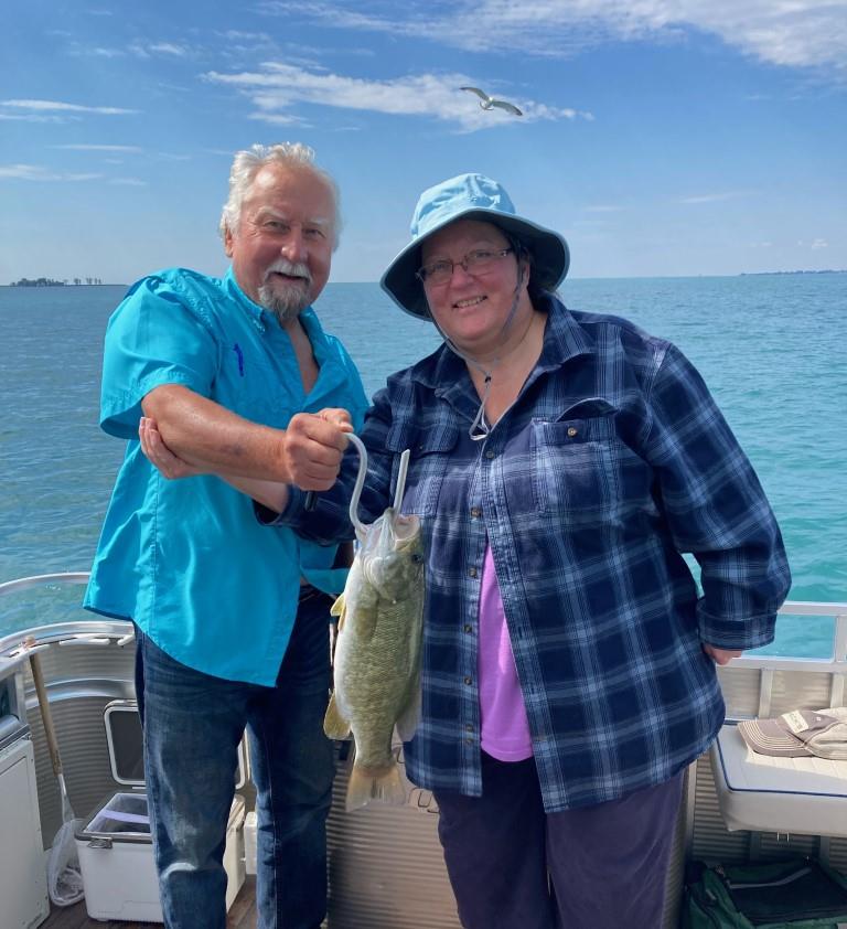 Two people on a boat holding two fish, blue water in the background. Sunny day.