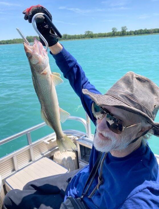 Man on a boat holds up a fish by its mouth, with blue water and sky in background.