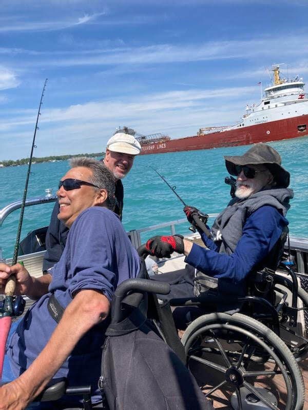 Three people fishing from a boat on blue water, a ship in the background. One person in a wheelchair.