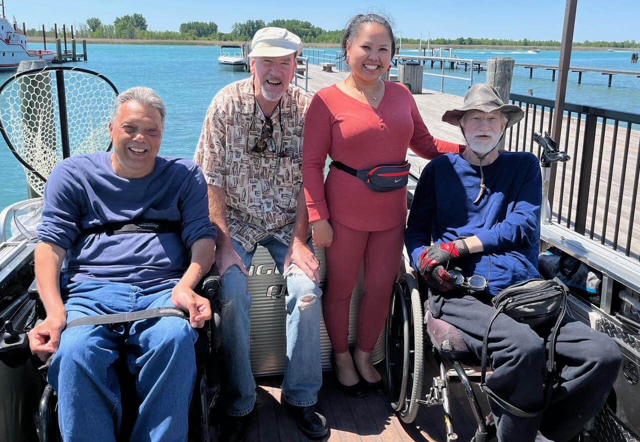 Four people on a dock with wheelchairs, sunny day, water in background. One woman, three men.