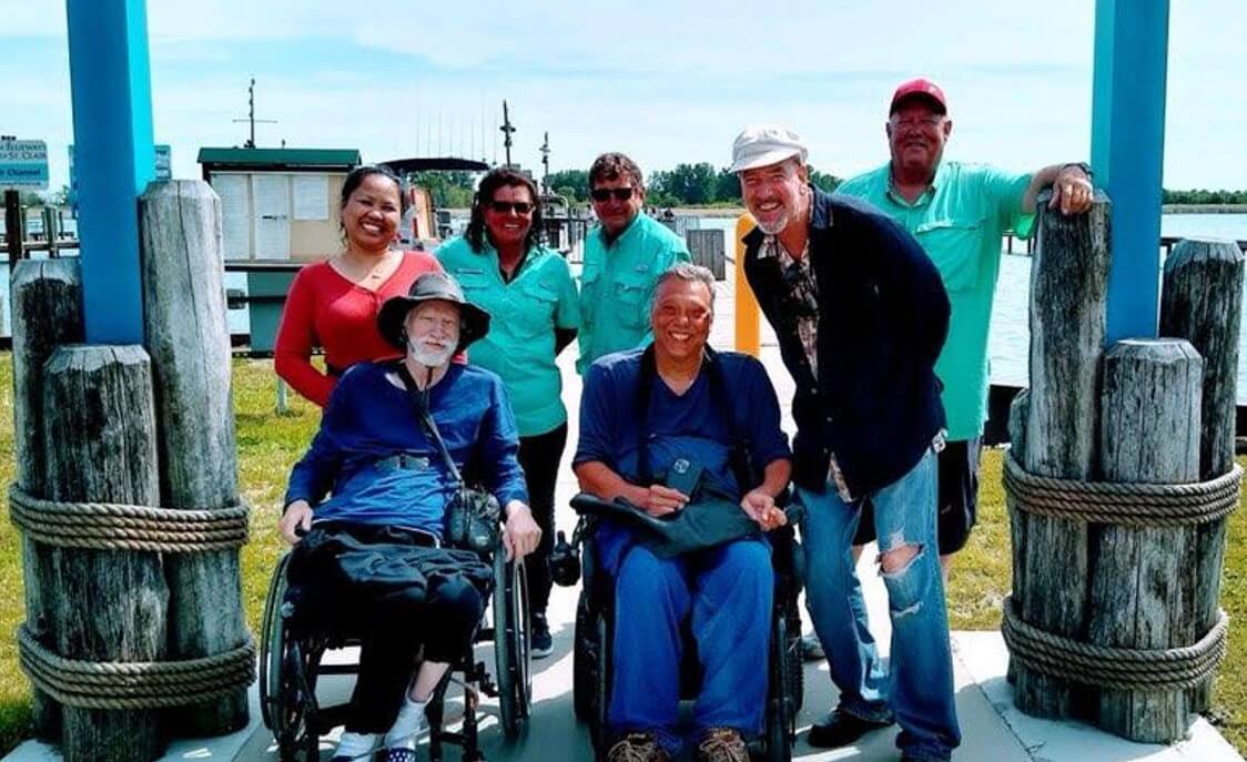 Group of people posing by a pier. Two are in wheelchairs. They are smiling and standing outside on a sunny day.