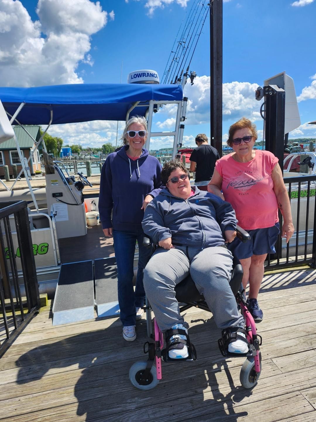 Three people on a dock. One in a wheelchair smiles. Others stand on either side. Boat with lift in the background. Blue sky.