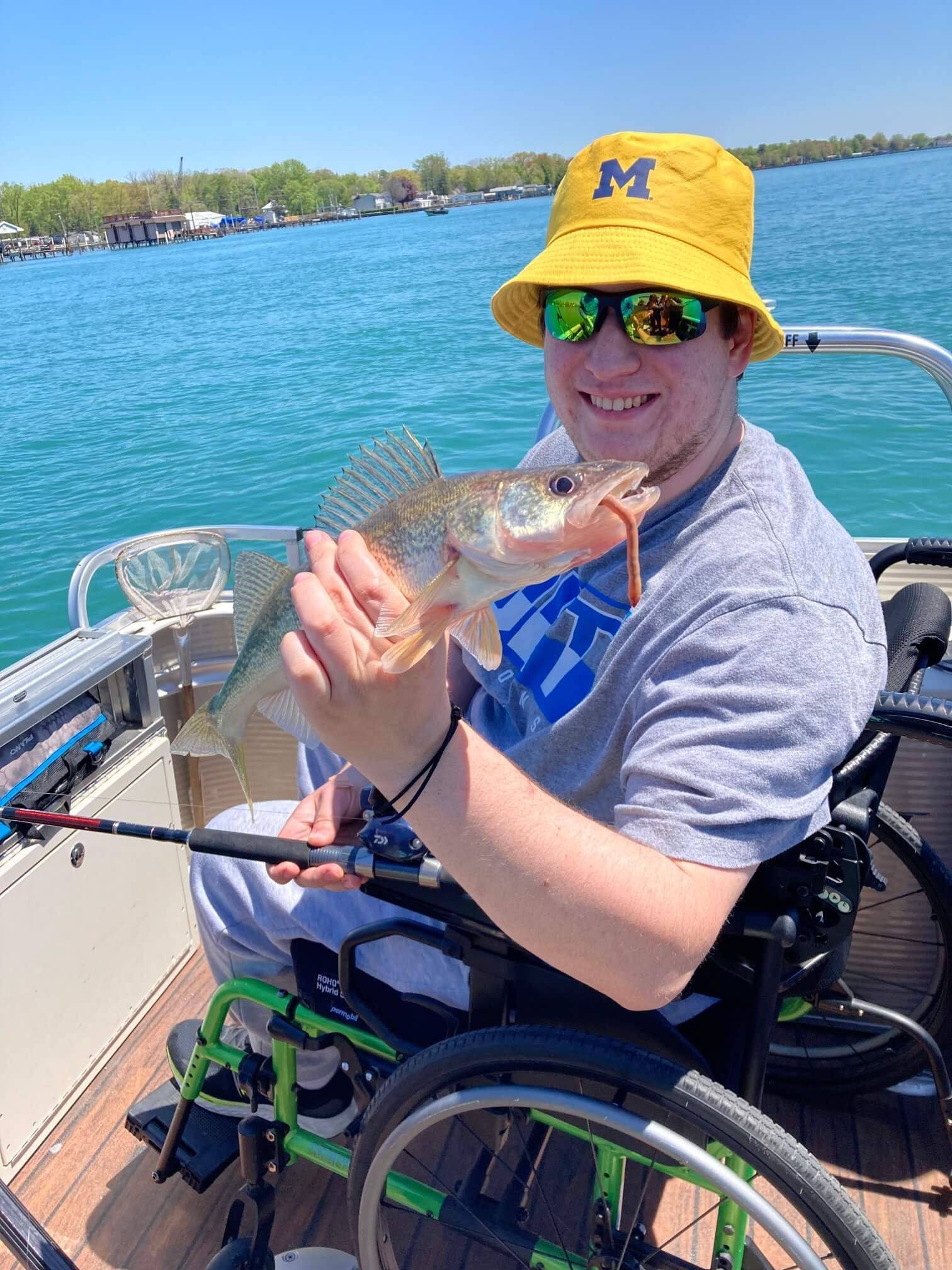 Person in wheelchair, holding a fish, smiles on a boat. Blue water, yellow hat, sunglasses.