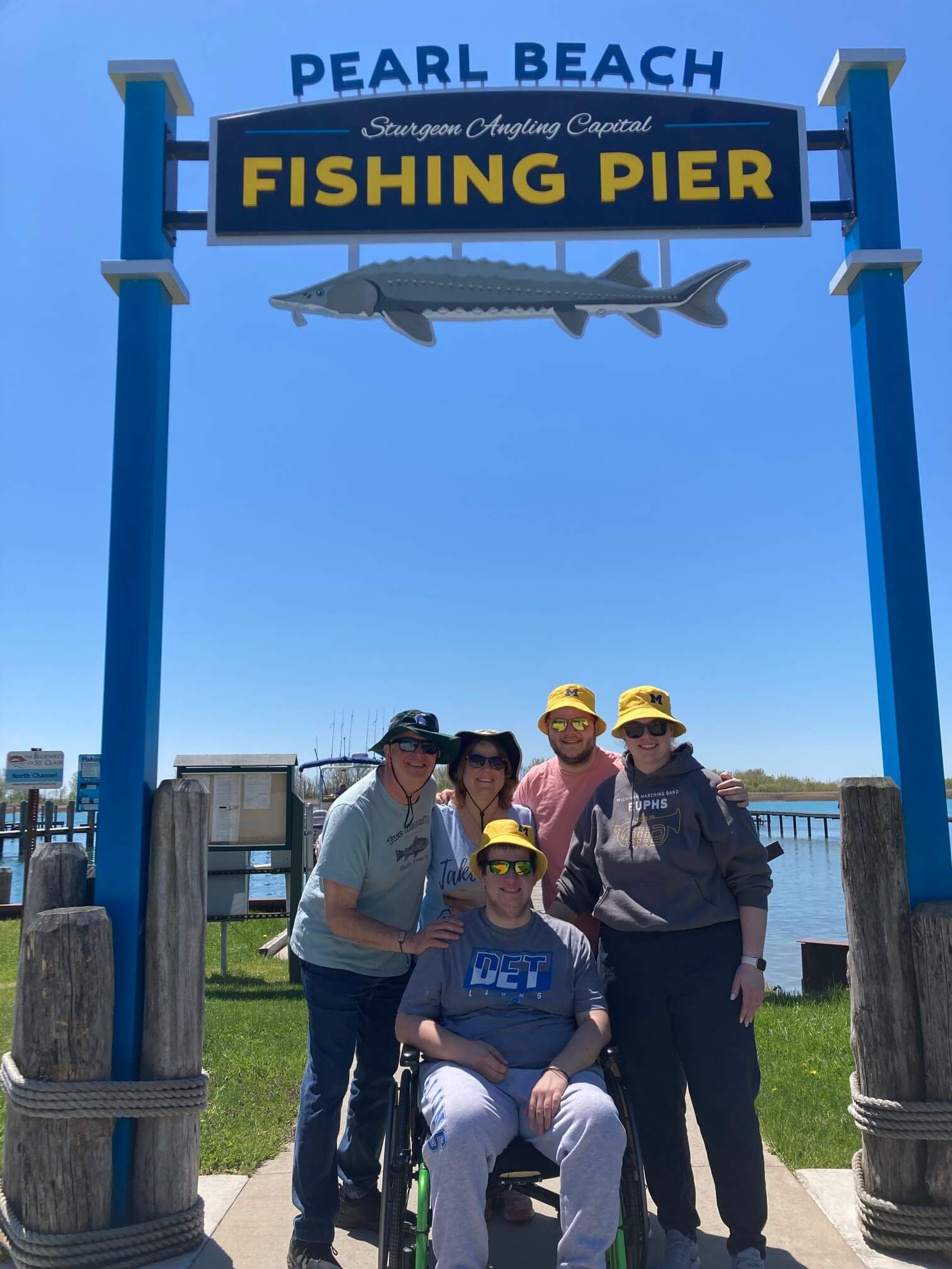 Group poses at Pearl Beach Fishing Pier sign under a blue sky, sturgeon statue on top.