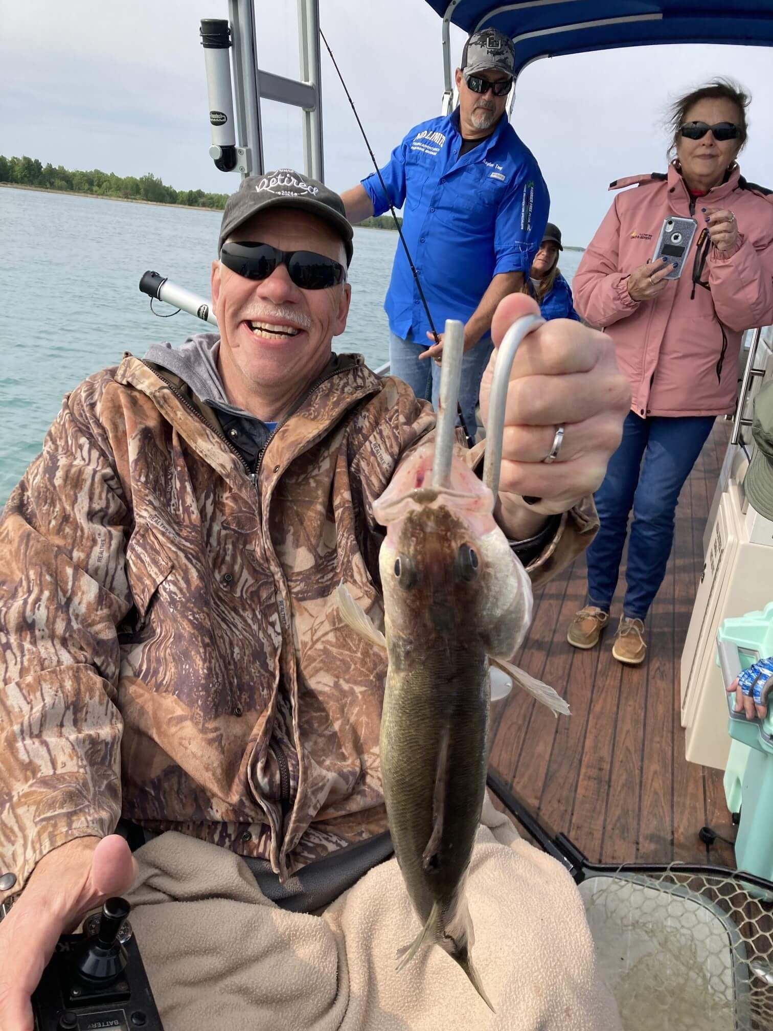 Man in camo jacket holding a fish on a boat. Other people visible, sunny day.