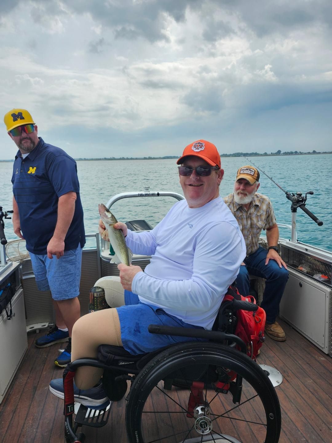 Man in wheelchair holding fish on boat, two others watching. Cloudy day, water.
