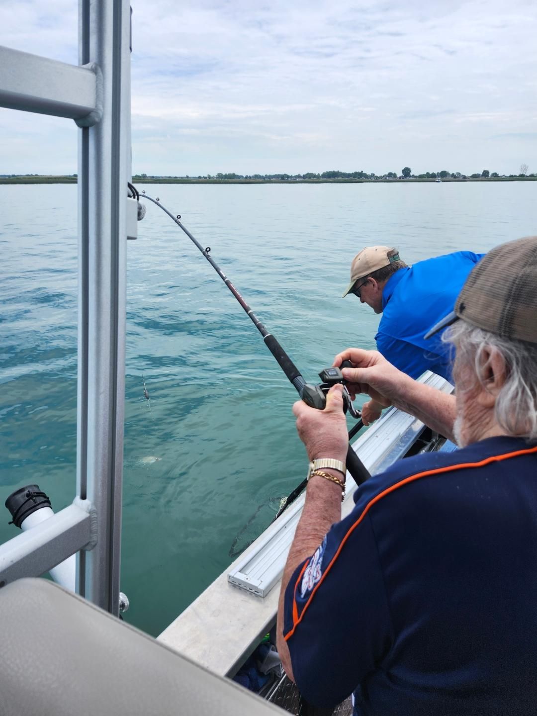 Two people fishing from a boat on blue water, cloudy sky, one reeling in a rod, other looking on.