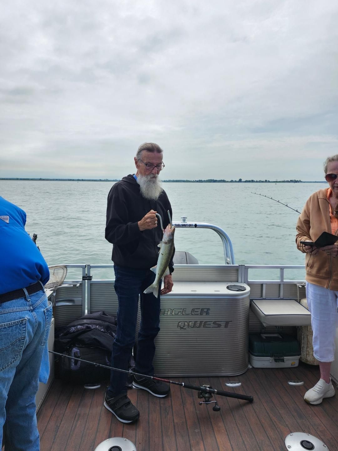 Man on a boat holds a fish. The setting is on a body of water with cloudy skies.
