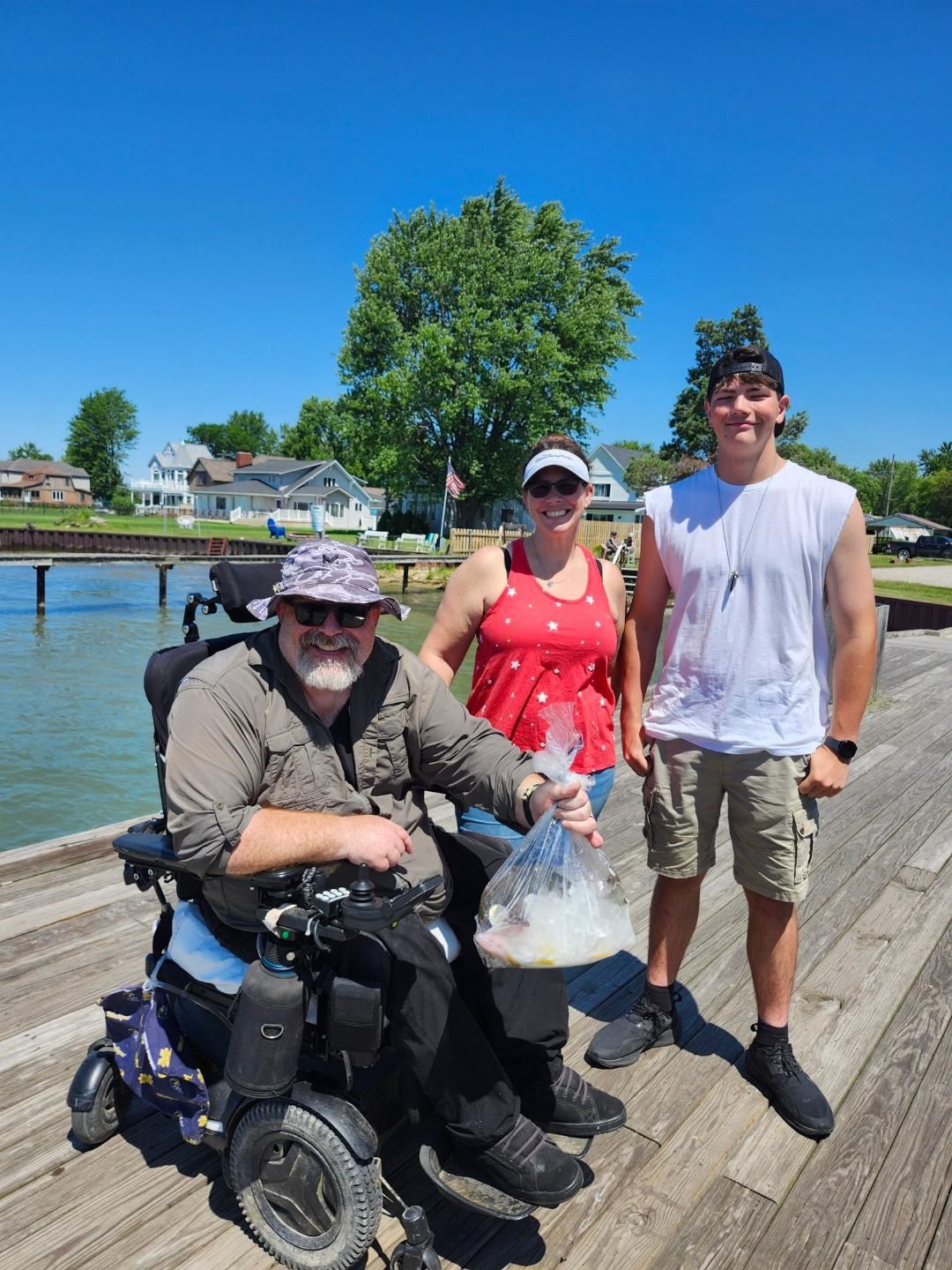 Three people on a wooden pier near water on a sunny day. Man in wheelchair holds bag. Woman and another man stand nearby.
