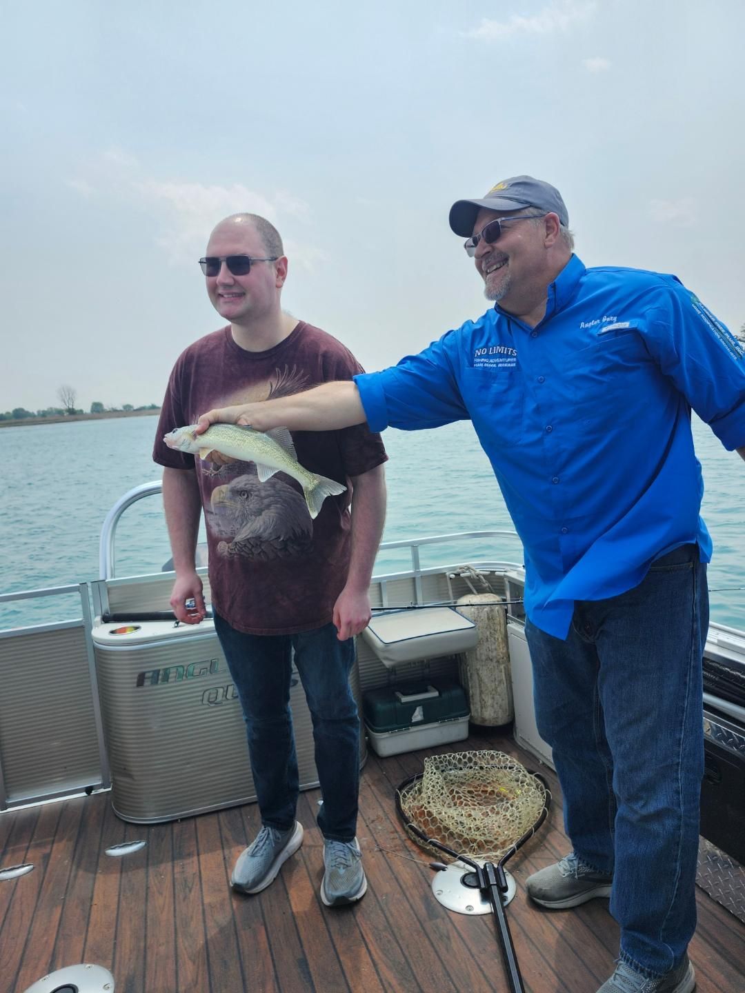 Two men on a boat; one holds up a fish, the other smiles. Blue water and sky.