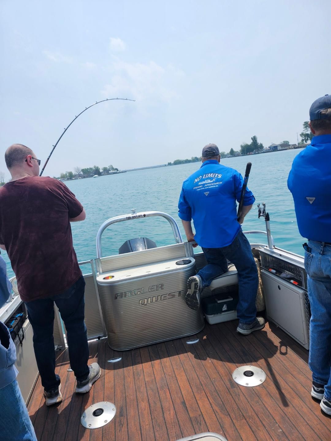 People fishing from a boat on a sunny day; one reeling in a fish. Water, blue shirts, rod.