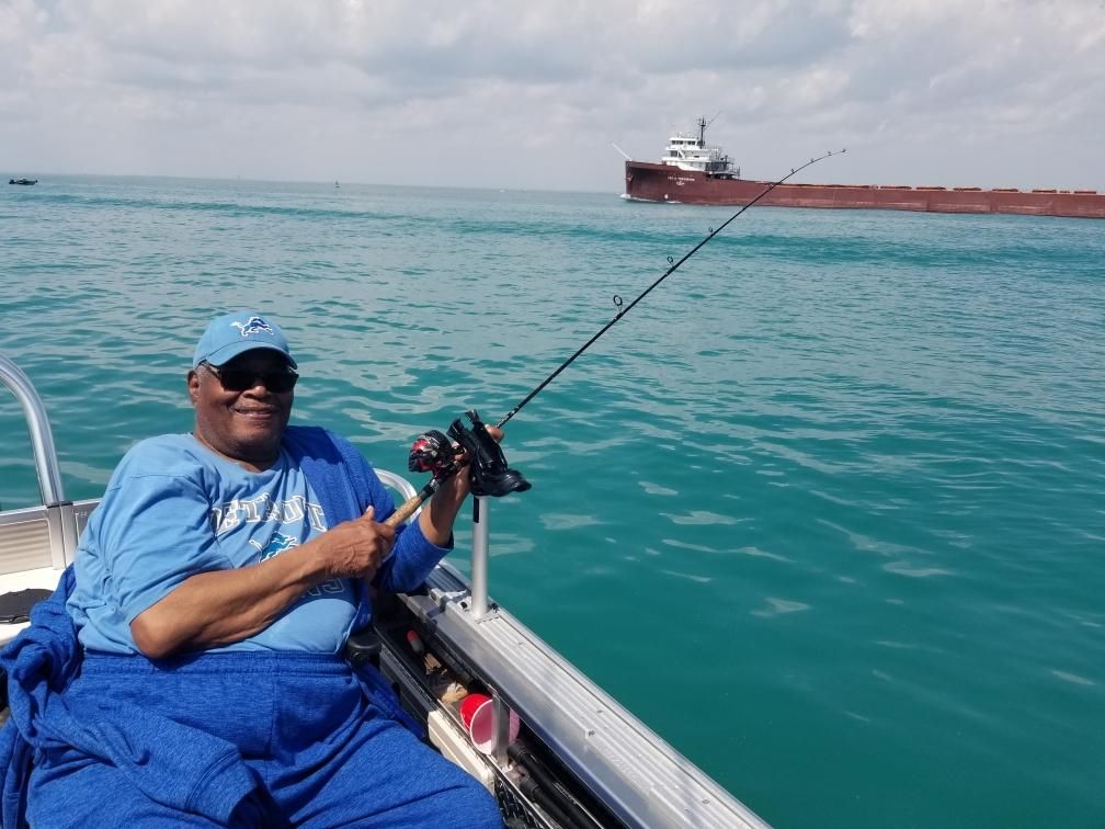 Person fishing on a boat, holding a rod. Calm, blue water with a large ship in the distance under a cloudy sky.