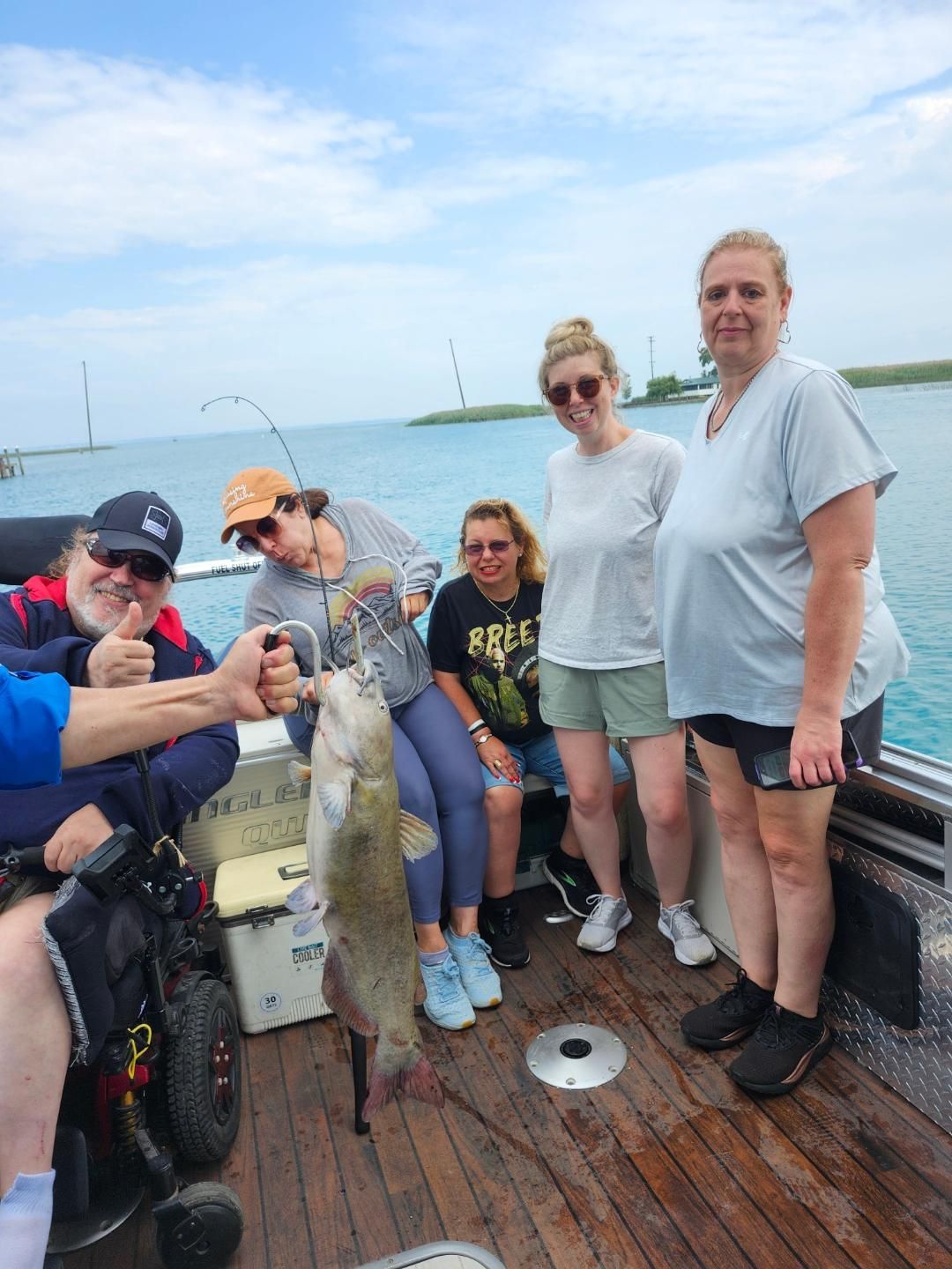 Group on a boat displays a caught fish. Person in wheelchair gives thumbs up. Cloudy sky, open water.