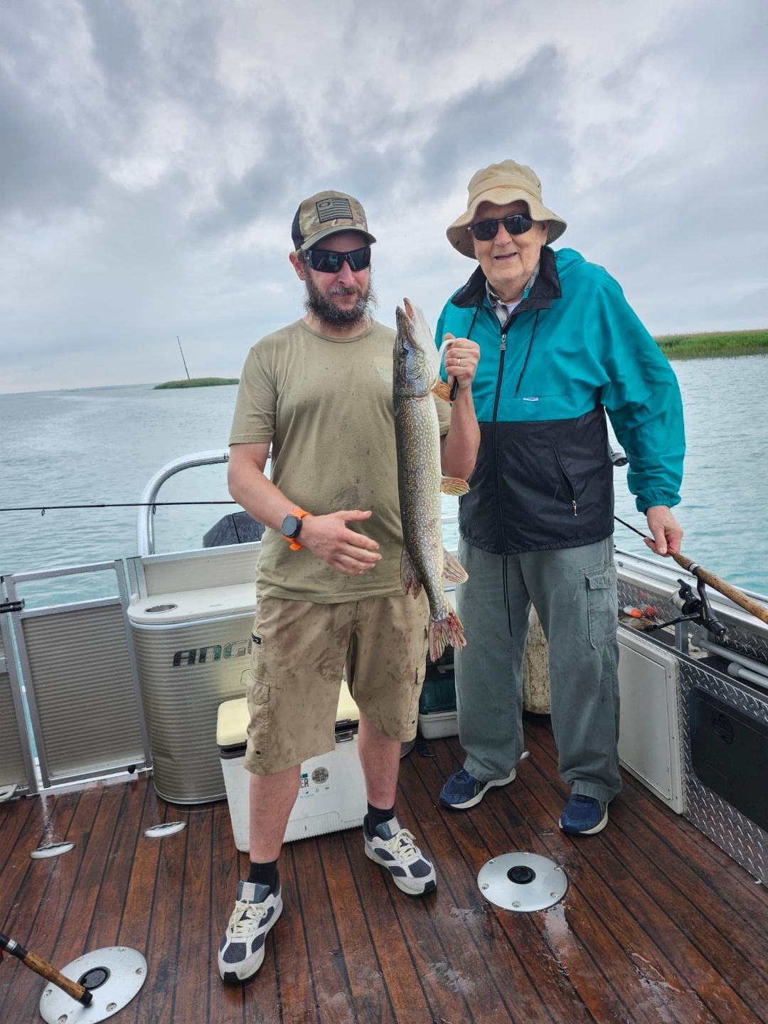 Two people on a boat display a fish. The man on the left holds it up; both smile. Cloudy sky, water backdrop.