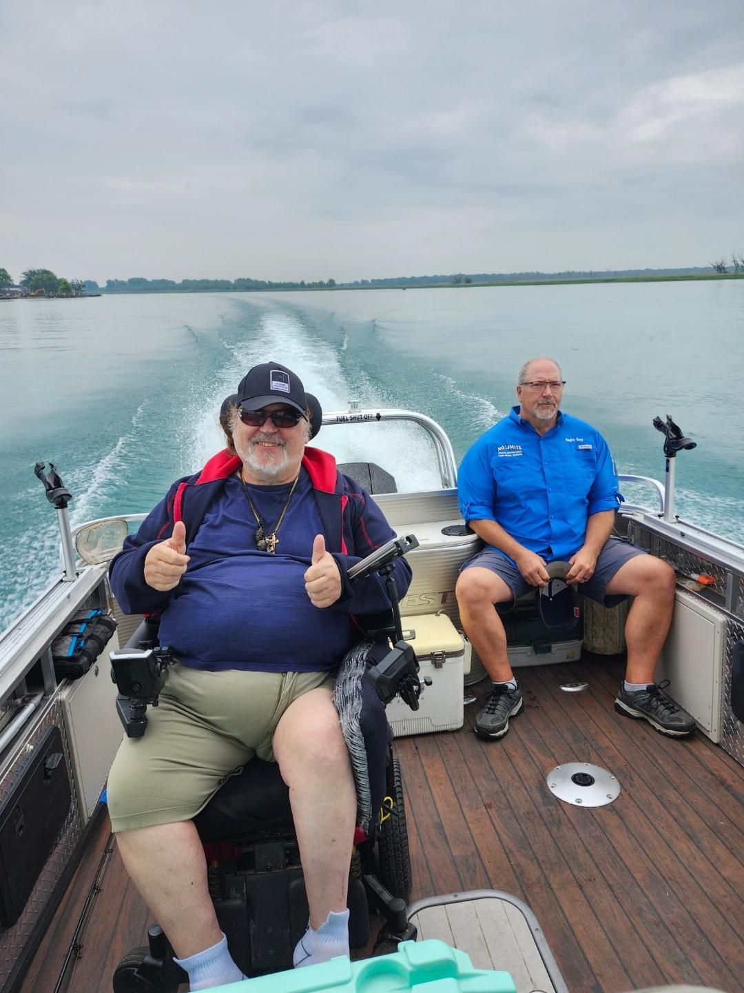 Two men on a boat, one in a wheelchair, thumbs up. They are on a lake with a blue sky.