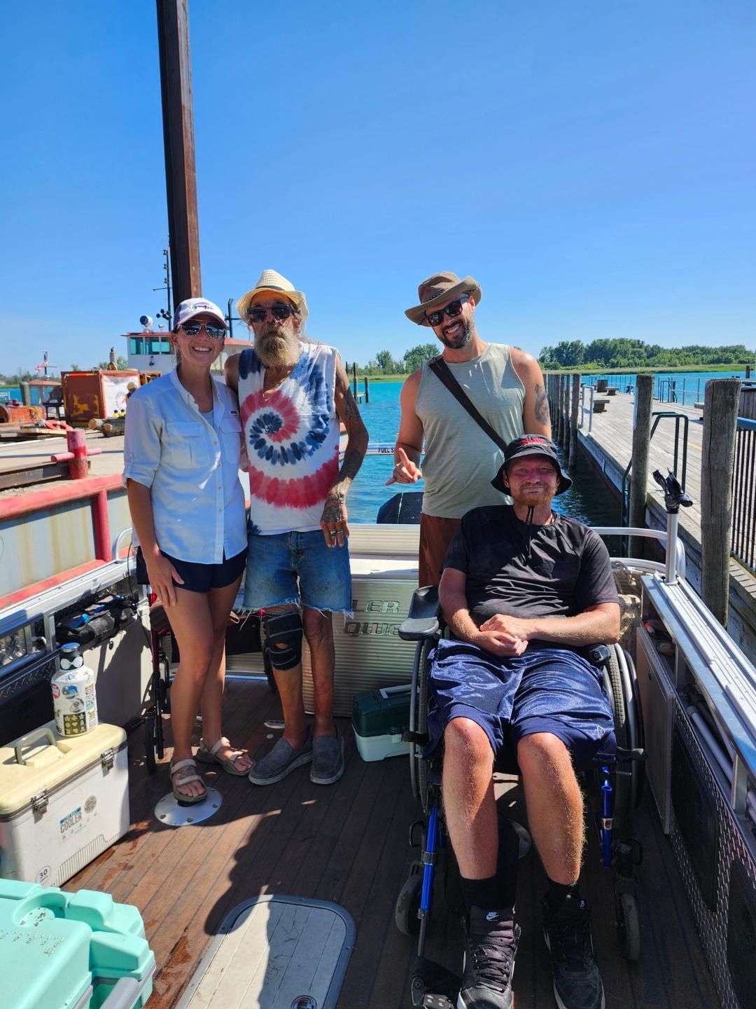 Group of four people on a boat, one in a wheelchair, near a dock on a sunny day.