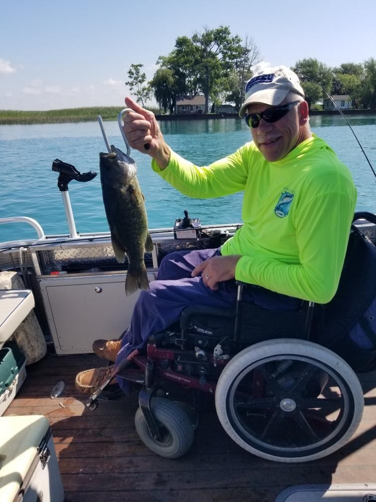 Man in wheelchair holds up a caught fish on a boat. Blue water and sky in the background.