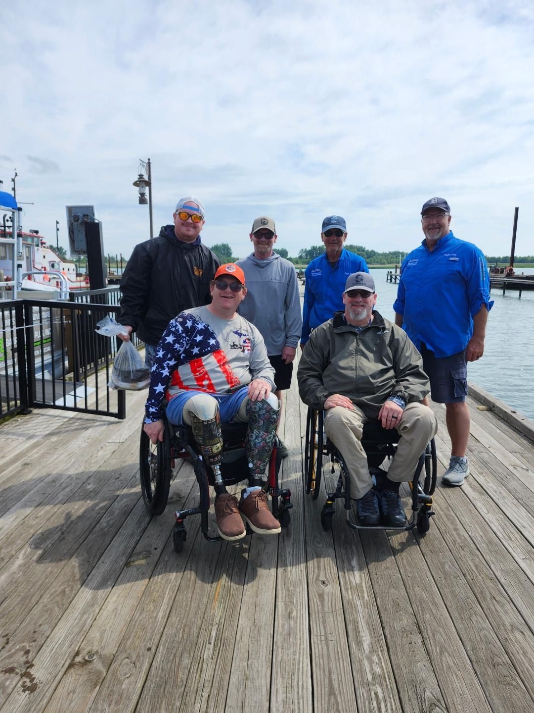 Group of six people on a wooden pier, two in wheelchairs. Cloudy sky, water, and boats in background.