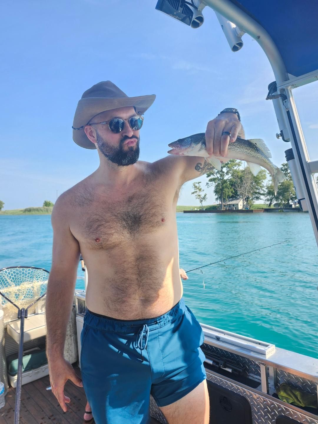 Man on a boat holding a fish, wearing a cowboy hat and sunglasses, on a sunny day.
