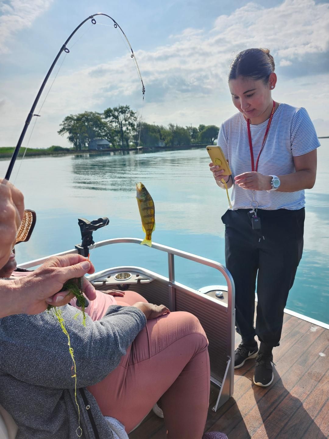 Person fishing on a boat; another person looks at a phone. A yellow fish is on the line. Blue water, sunny sky.