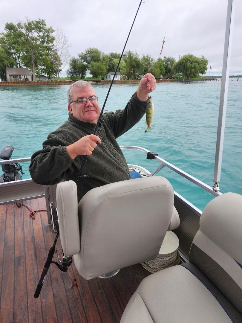 Man on a boat, holding a small fish up in the air. Turquoise water, green trees in background.