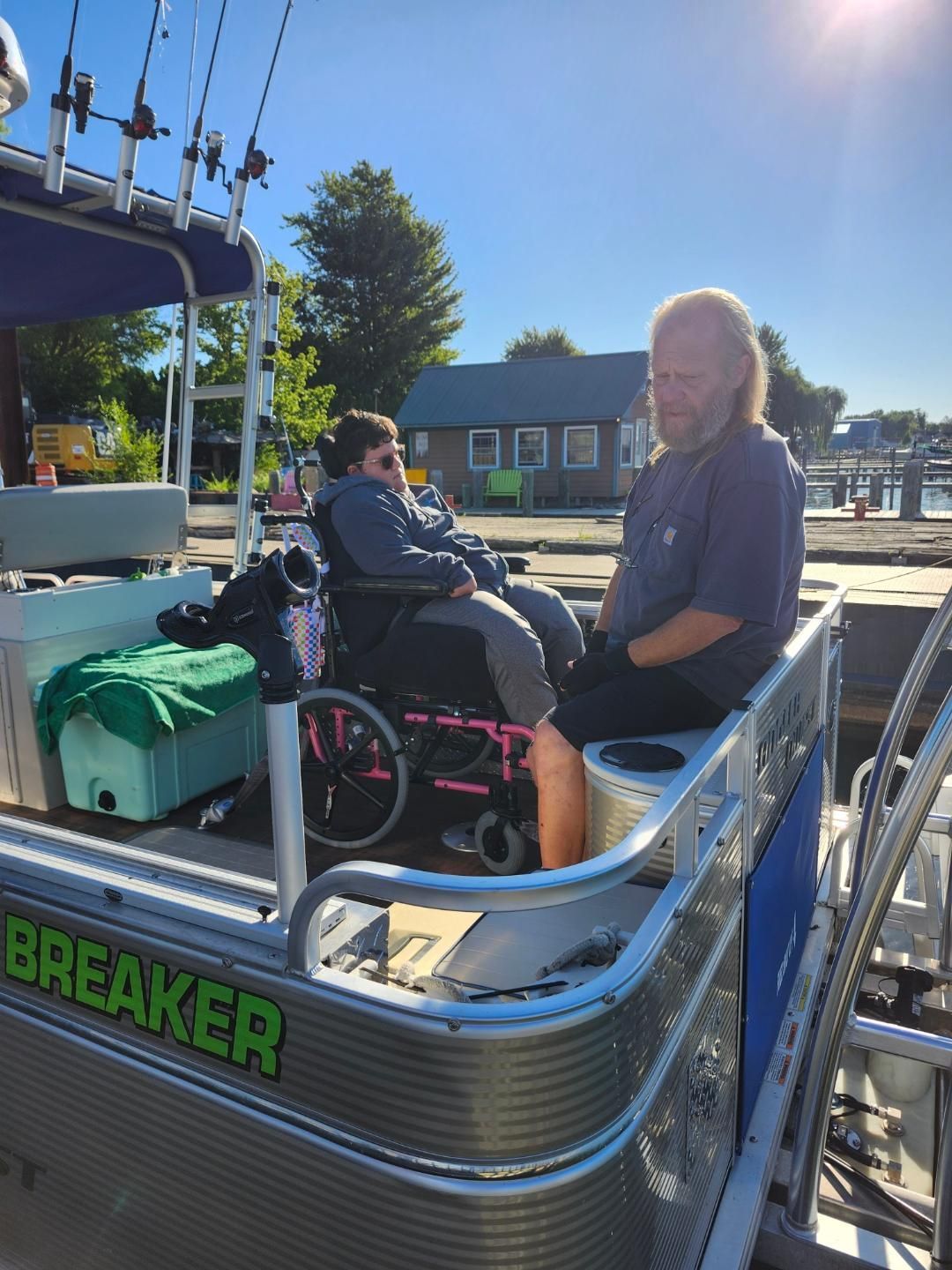 Man and person in wheelchair on a boat. The boat has fishing rods. Outdoors on a sunny day.