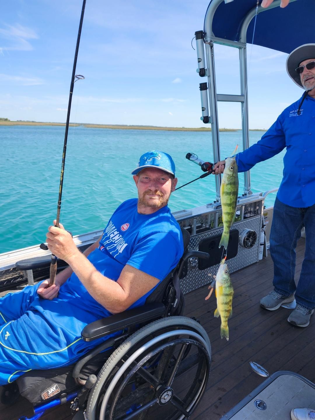 Man in wheelchair fishing on a boat; another man holds up two yellow perch.