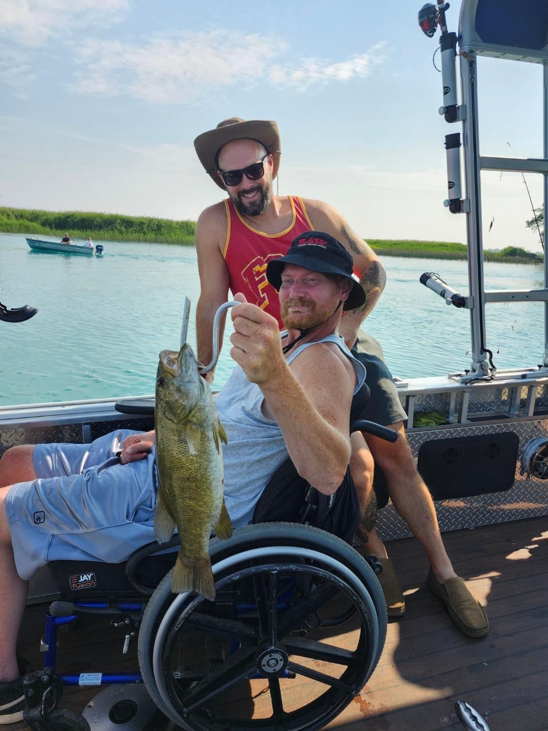 Man in wheelchair on a boat holding a fish; another man behind him smiling, on a lake.