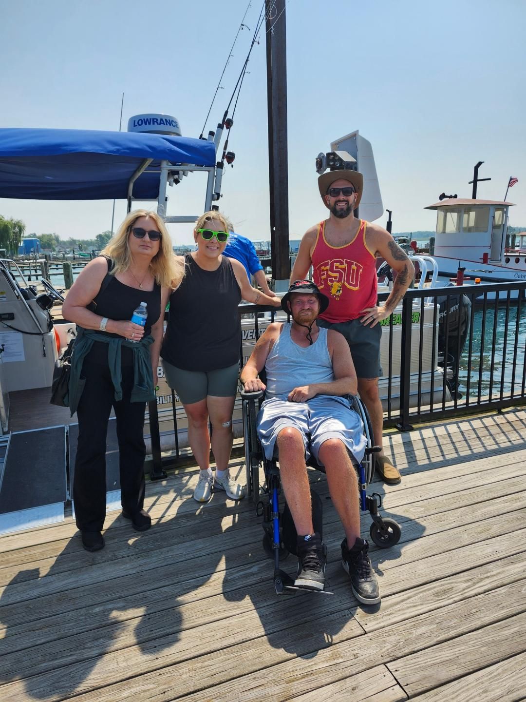 Four people pose on a dock by a boat. One sits in a wheelchair; others stand. Sunny day.