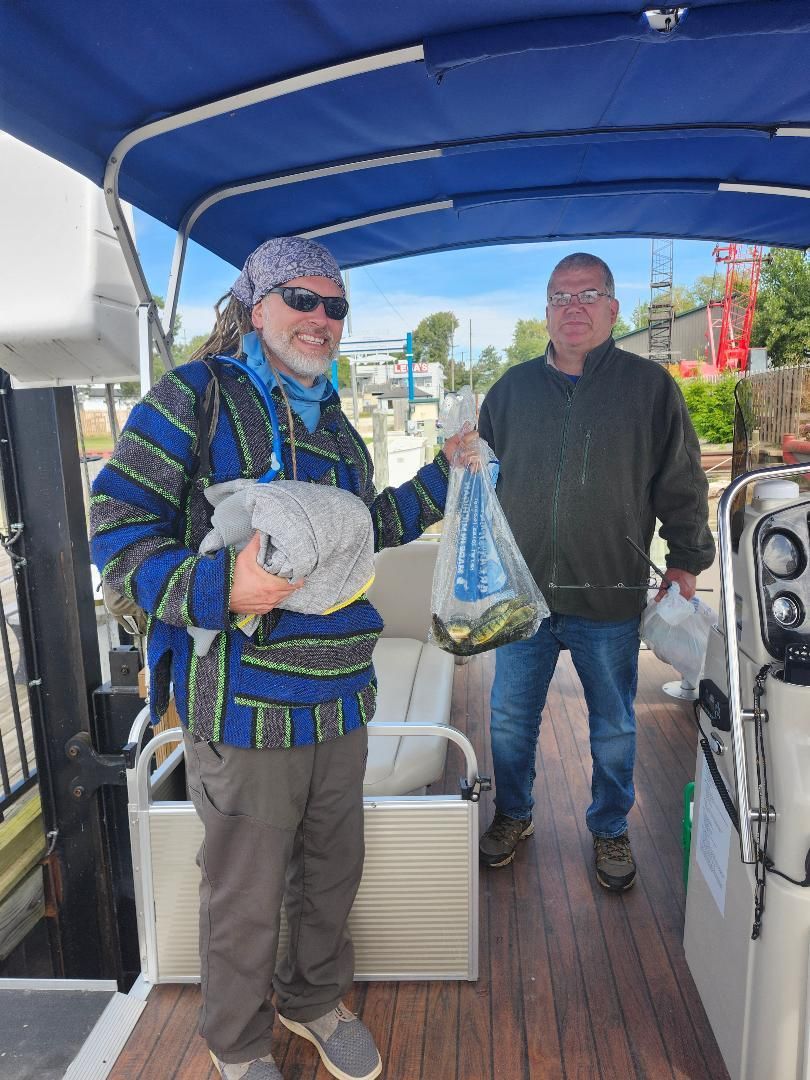 Two men on a boat, holding bags. One wears a patterned jacket, the other a green sweater. Outdoors, sunny.
