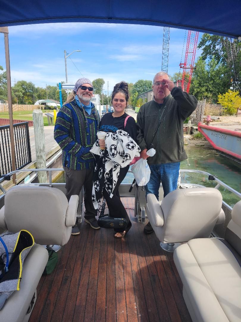 Three people on a boat near a dock; woman holding a blanket, man waving, another man smiling.