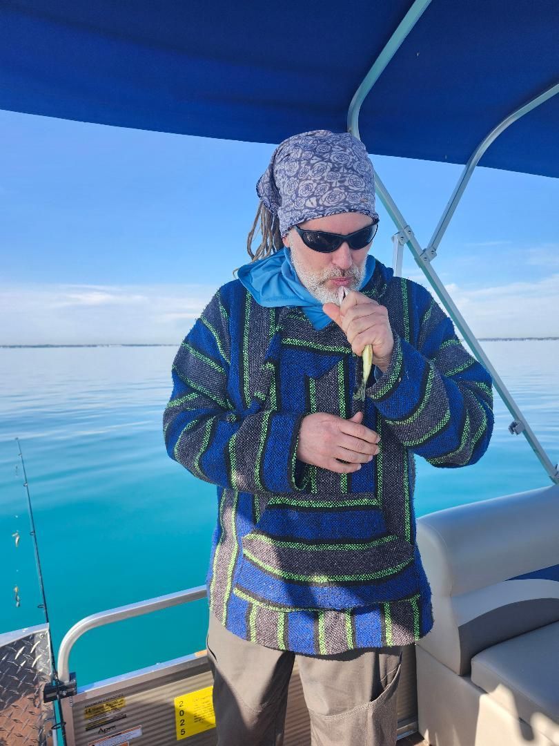 Man on a boat, wearing sunglasses and a patterned shirt, holding a small fish, near water.