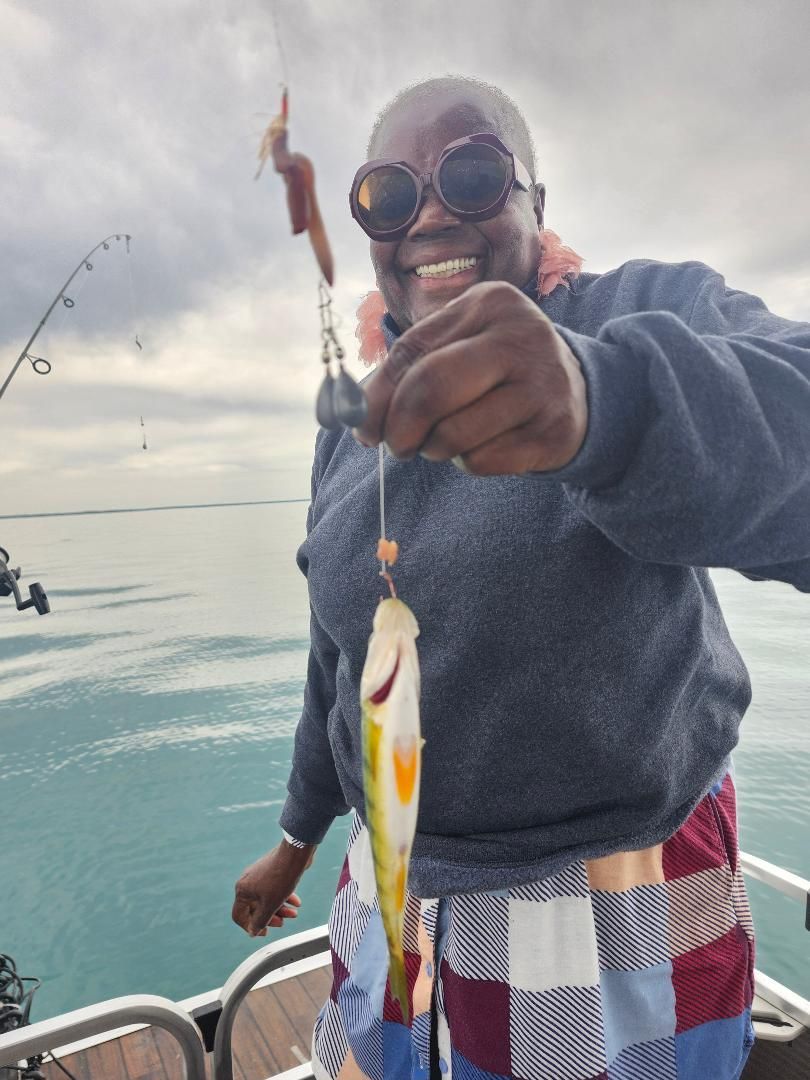 Smiling person on a boat, holding up a small yellow fish with fishing line. Gray water and cloudy sky.