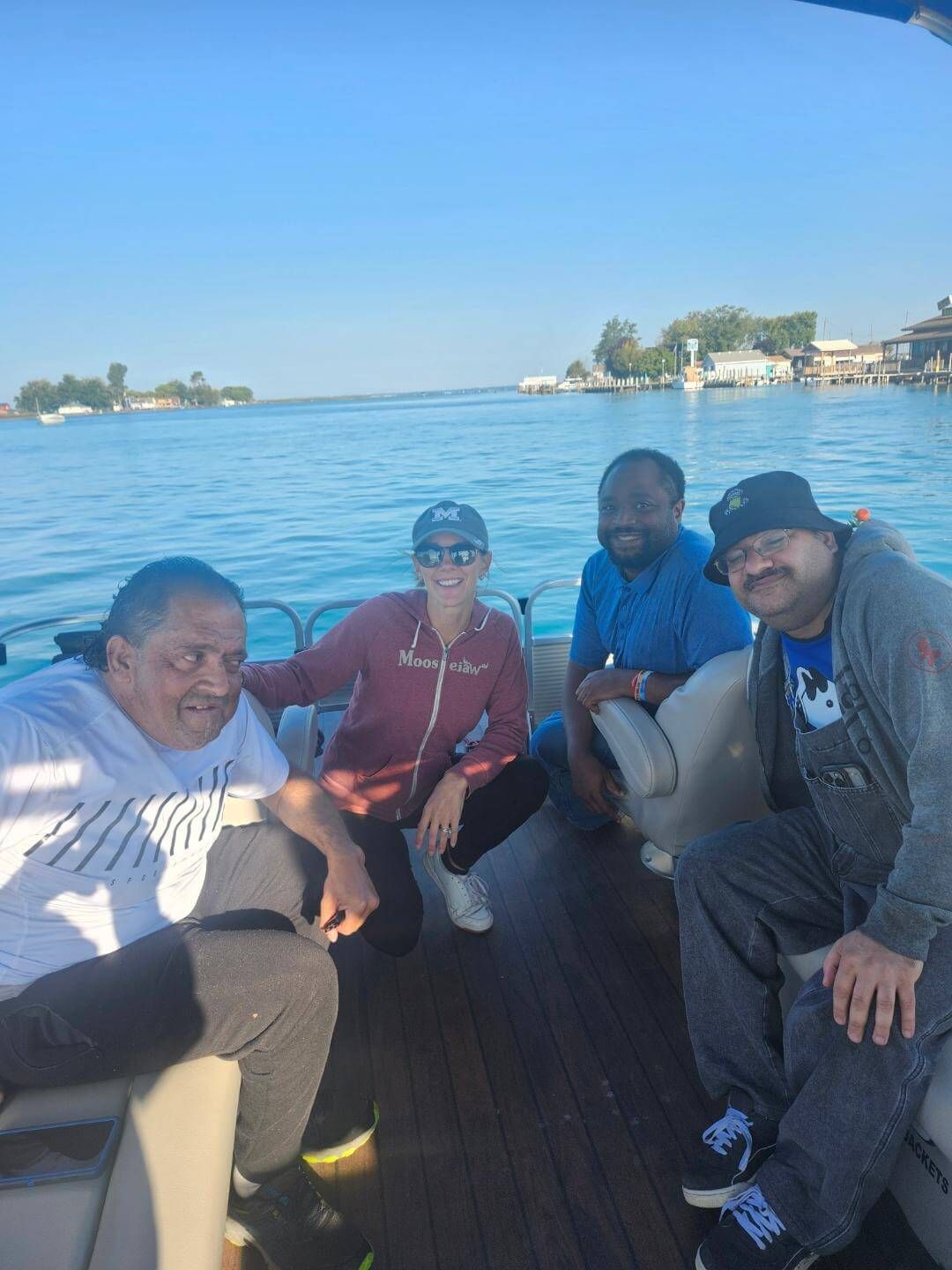 Four people smile on a boat. The sunny scene has a blue sky and water. Buildings line the shore in the background.