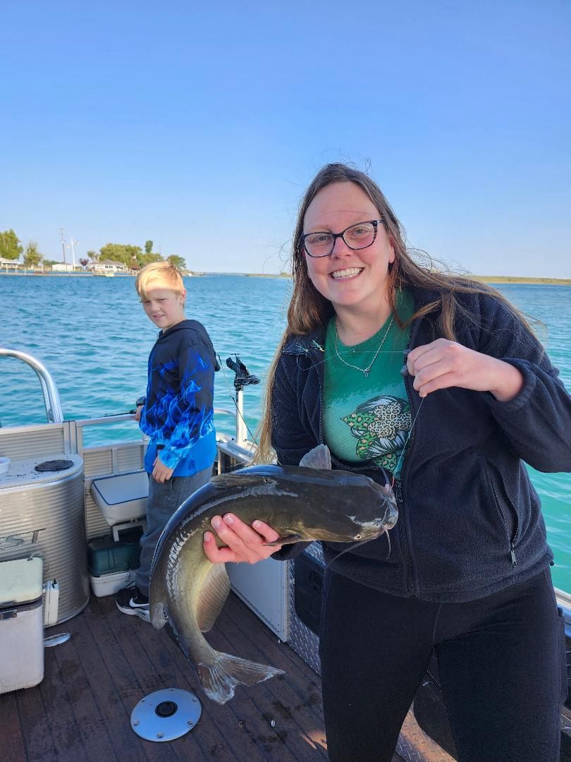 Woman on boat holding a large fish, smiling. Boy fishing in the background, blue water and sky.