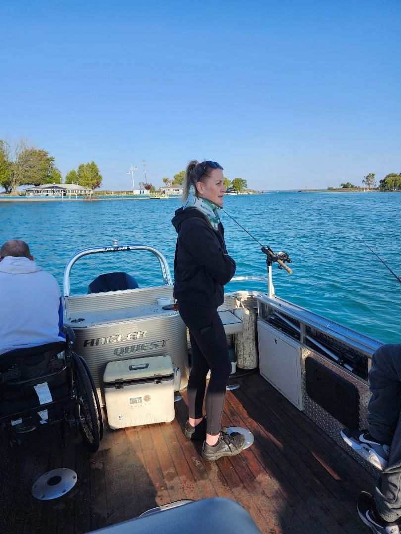 Woman standing on a boat fishing. Calm water, clear sky, and shoreline in the background.