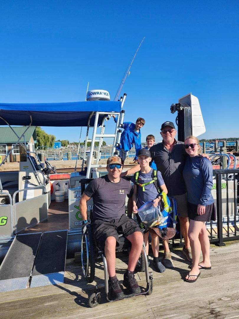 Family on a boat dock, some smiling, one in a wheelchair, fishing pole in background, sunny day.