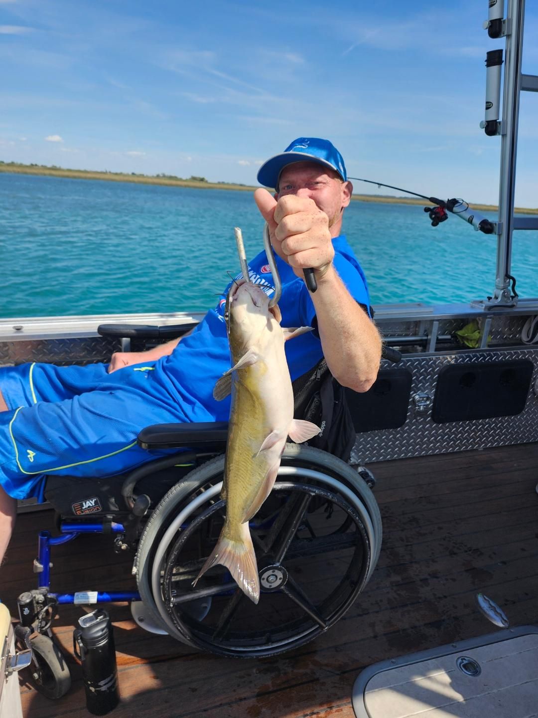 Man in wheelchair on a boat holding up a catfish, giving a thumbs-up. Blue water, sunny sky.