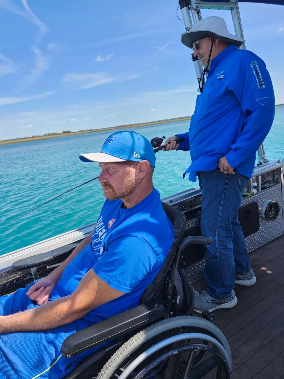 Man in wheelchair fishing on a boat; another man assists. Clear blue water and sky.
