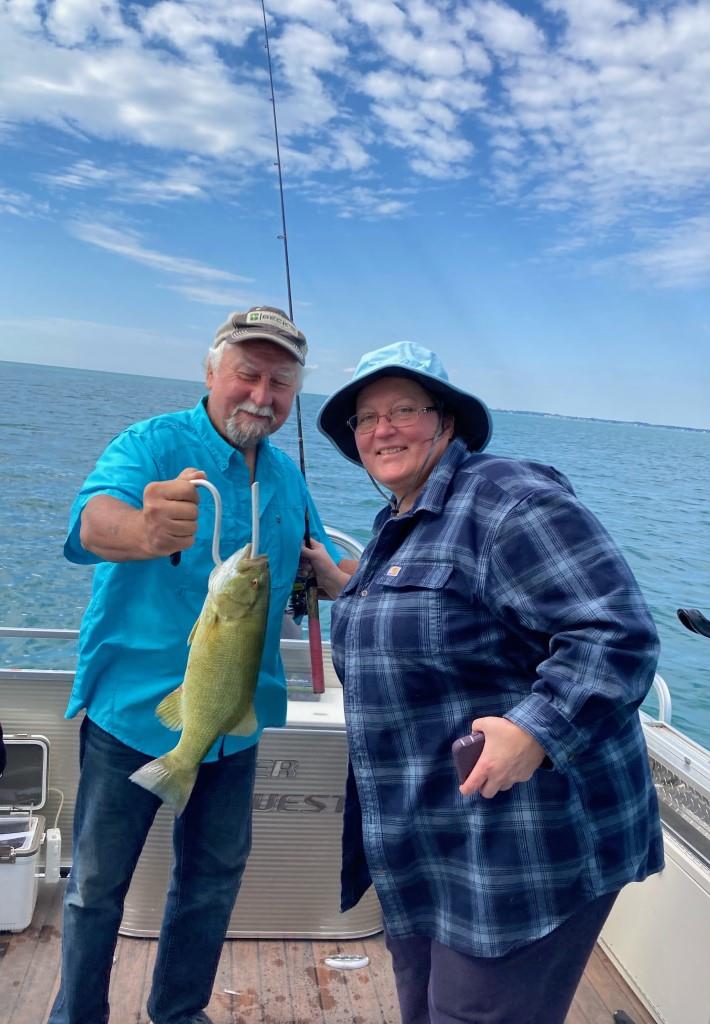 Two people on a boat display a caught fish. The man holds the fish. Overcast sky, water visible.