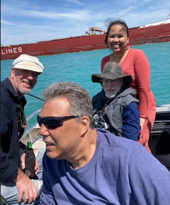 Four people on a boat near a large red ship, enjoying the turquoise water on a sunny day.