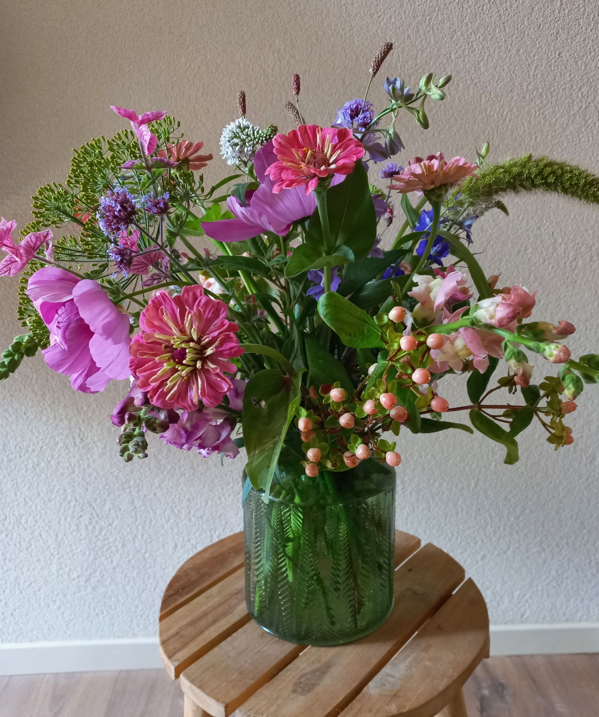 Boeket roze, paarse en blauwe bloemen in een groene glazen vaas op een houten krukje.